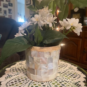 Può includere: Un vaso di ceramica bianca con un bordo dorato e un motivo a mosaico di piastrelle bianche e beige. Il vaso è pieno di fiori bianchi e foglie verdi.