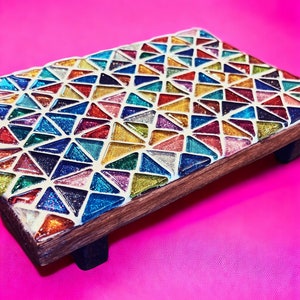 May include: A colourful mosaic tray with a geometric pattern of triangles in shades of red, orange, yellow, green, blue, and purple. The tray is made of wood and has a glossy finish. It is sitting on a pink surface.