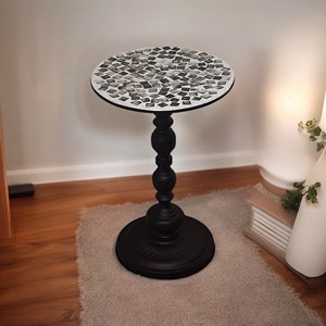 Op de afbeelding: Een zwarte bijzettafel met een rond mozaïek tafelblad. De mozaïek is gemaakt van kleine, grijze en witte tegels. De tafel heeft een zwarte, sierlijke basis.
