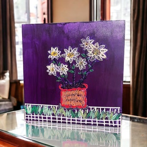 Op de afbeelding: Een paars canvas schilderij van witte papieren bloemen in een rode pot met groene bladeren. De bloemen zijn gemaakt van gekruld papier en zijn gerangschikt in een boeket. Het schilderij staat op een houten tafel.