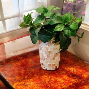 Op de afbeelding: Een decoratieve vaas met een mozaïek schelpontwerp, gevuld met levendige groene planten. De vaas staat op een rood-oranje tafel bij een raam.