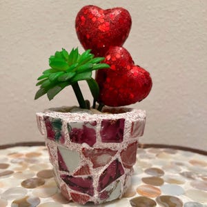 May include: A small, pink mosaic planter with a green succulent plant and two red glitter hearts.