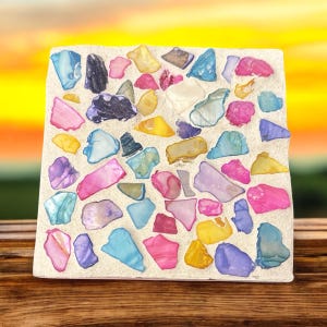 Op de afbeelding: Een vierkante onderzetter van wit beton met een mozaïek van kleurrijke glazen stukjes die in het oppervlak zijn ingebed.