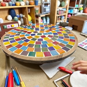 Könnte beinhalten: Ein runder Mosaiktisch mit Holzrahmen. Die Tischplatte ist mit bunten quadratischen Fliesen in Rot-, Orange-, Gelb-, Grün-, Blau- und Lilatönen bedeckt. Ein dekoratives Element.