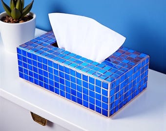 May include: A rectangular tissue box covered in small, square, blue mosaic tiles. White tissues are visible, emerging from the top opening. The box sits on a white surface, with a small potted plant in the background.