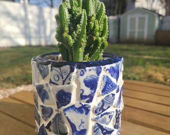 Può includere: Una pianta di cactus in un vaso di mosaico blu e bianco. Il vaso è fatto di pezzi di porcellana rotti, creando un aspetto unico e decorativo.
