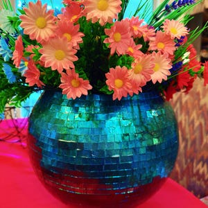 Puede incluir: Un jarrón esférico con mosaicos azules y verdes contiene un arreglo de flores artificiales. Las flores son principalmente margaritas de color melocotón con centros amarillos, complementadas con follaje verde y toques de azul y rojo.