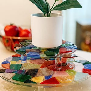 Può includere: Un vassoio di vetro a due livelli con un design a mosaico di pezzi di vetro colorati. Un vaso di ceramica bianco con una pianta verde è posto sopra. Il vassoio è sostenuto da gambe cilindriche trasparenti. I pomodori rossi sono visibili sullo sfondo.