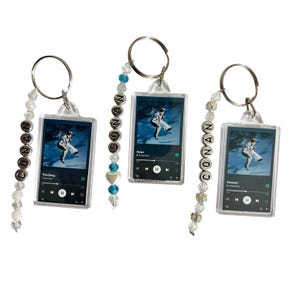 Puede incluir: Tres llaveros de acrílico transparente con una interfaz de reproductor de música y una foto de una persona. Cada llavero tiene un llavero plateado y un dije de cuentas con el nombre "CONAN". Los llaveros tienen diferentes títulos de canciones.