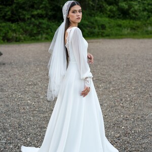 May include: A bride wearing a white wedding dress with long sleeves and a veil. The dress has a lace bodice and a flowing skirt. The bride is standing on a gravel path.