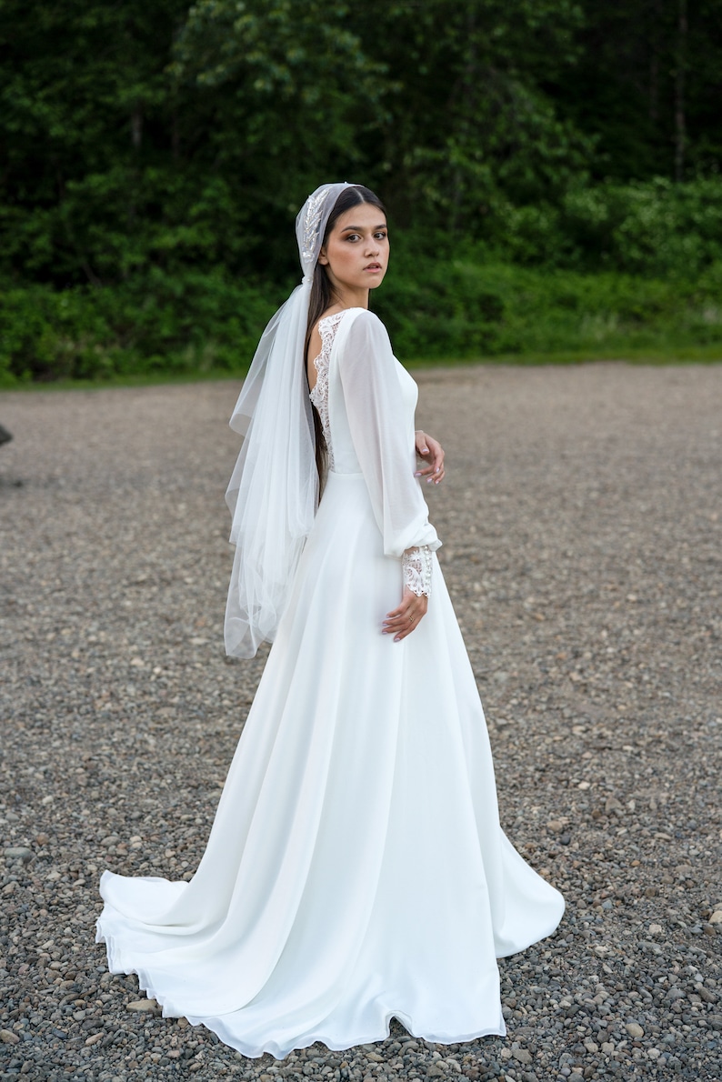 May include: A bride wearing a white wedding dress with long sleeves and a veil. The dress has a lace bodice and a flowing skirt. The bride is standing on a gravel path.