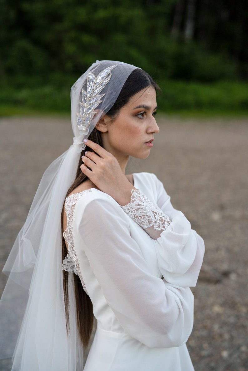 May include: A bride wearing a white wedding dress with lace detailing on the sleeves and back. She is wearing a veil with a jeweled headpiece.