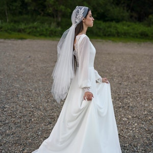 May include: A bride wearing a white wedding dress with long sleeves and a veil. The dress has a lace back and the veil is decorated with a silver floral design. The bride is standing on a gravel path.