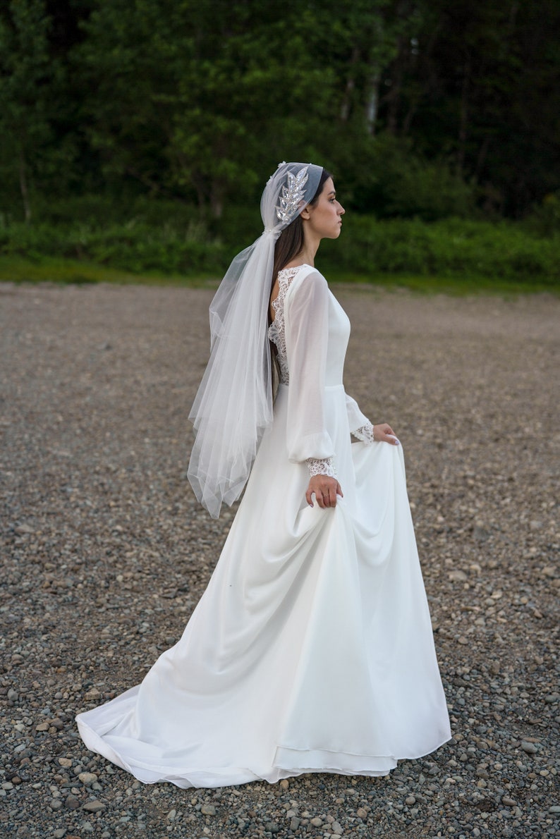 May include: A bride wearing a white wedding dress with long sleeves and a veil. The dress has a lace back and the veil is decorated with a silver floral design. The bride is standing on a gravel path.