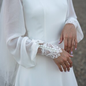 May include: A white wedding dress with long sleeves and a lace detail on the cuffs. The dress has a simple, elegant design.