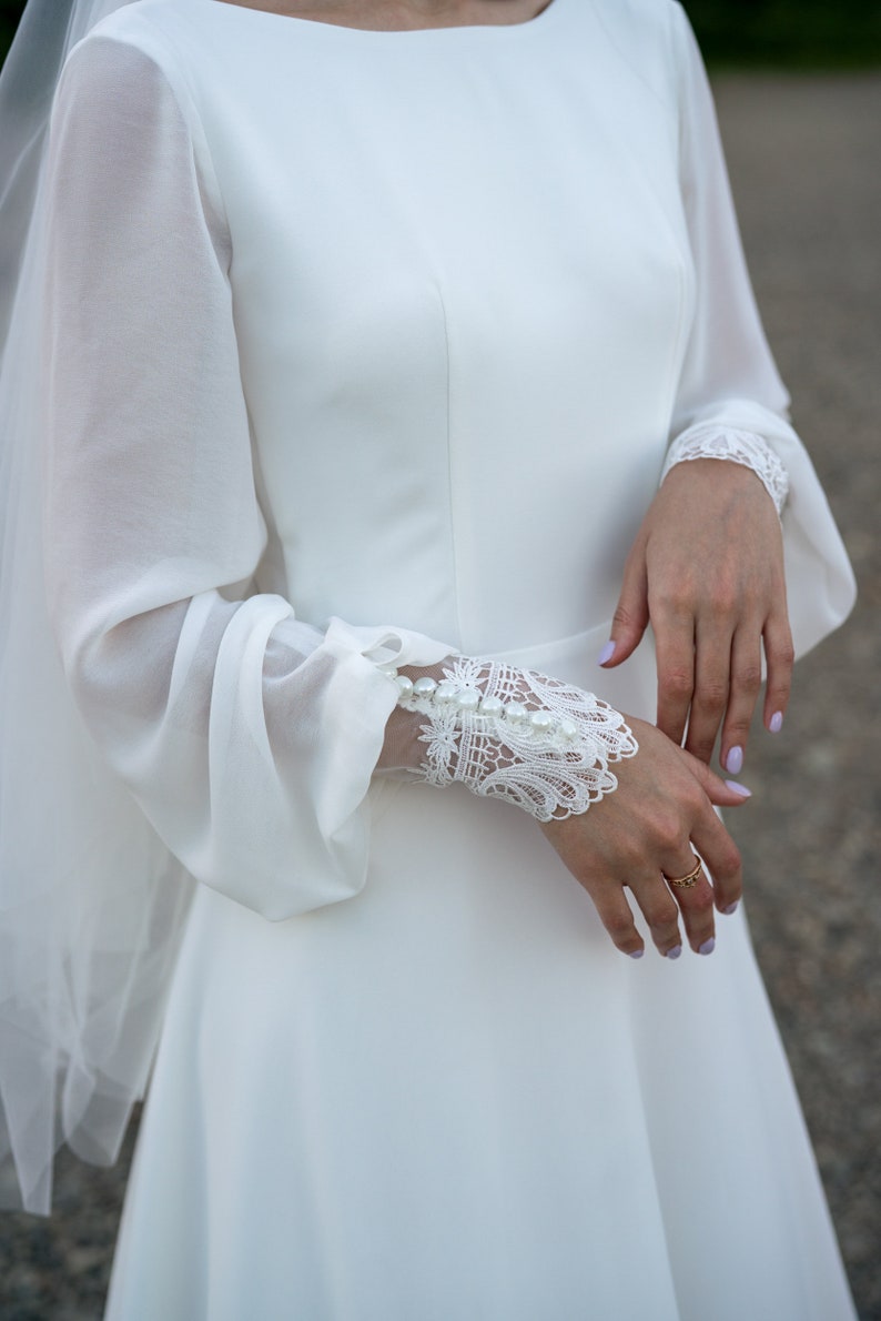 May include: A white wedding dress with long sleeves and a lace detail on the cuffs. The dress has a simple, elegant design.