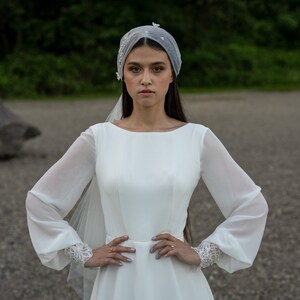 May include: A bride wearing a white wedding dress with long sleeves and a veil. The dress has a fitted bodice and a flowing skirt. The bride is standing in front of a green background.