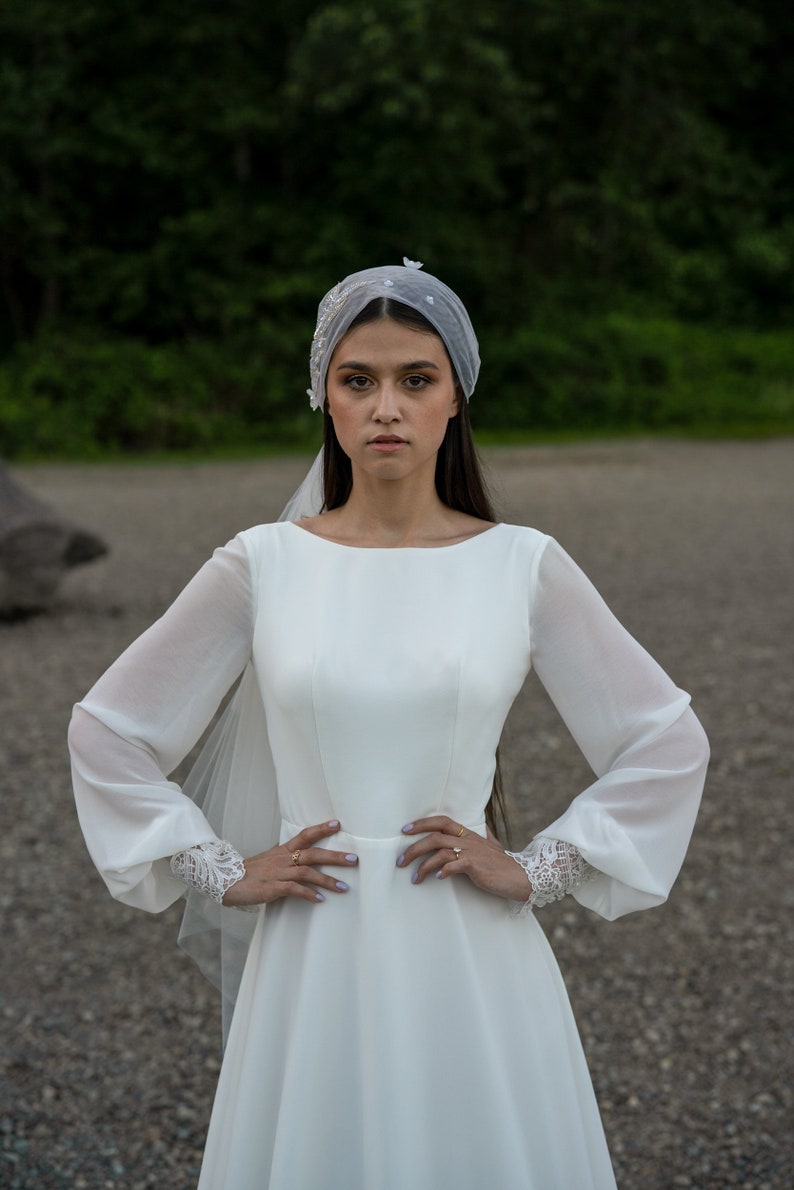 May include: A bride wearing a white wedding dress with long sleeves and a veil. The dress has a fitted bodice and a flowing skirt. The bride is standing in front of a green background.