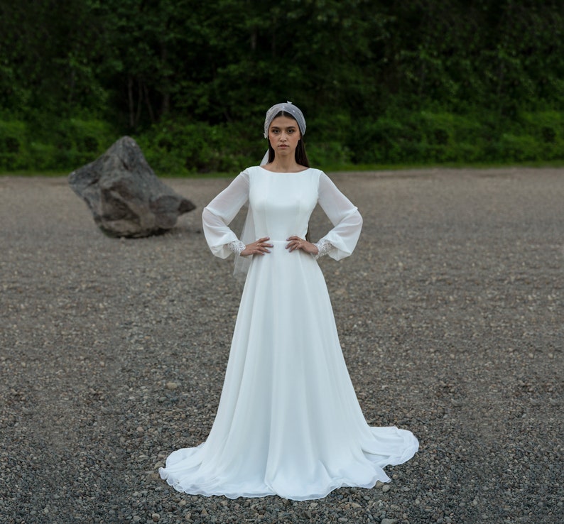 May include: A white wedding dress with long sleeves and a flowing skirt. The dress has a simple, elegant design and is perfect for a bride who wants a classic look.