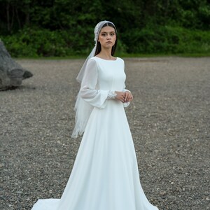 May include: A white wedding dress with long sleeves and a flowing skirt. The dress has a simple, elegant design and is perfect for a bride who wants a classic look.