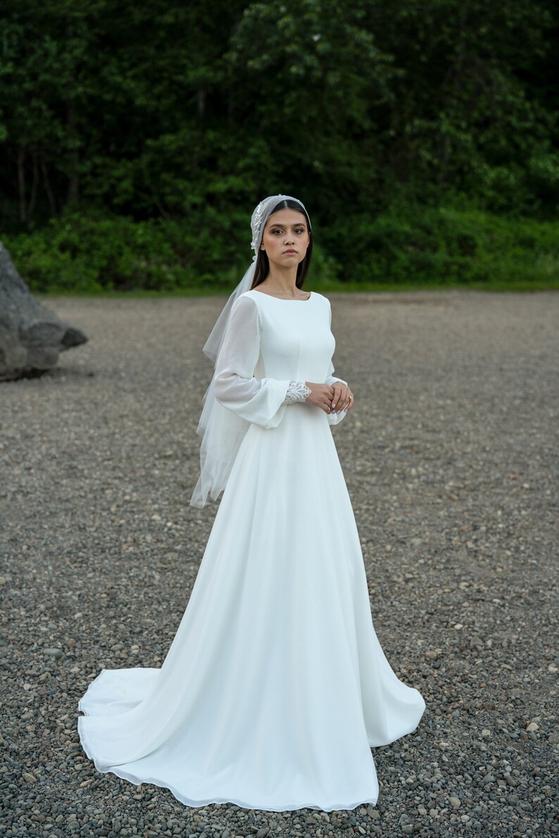 May include: A white wedding dress with long sleeves and a flowing skirt. The dress has a simple, elegant design and is perfect for a bride who wants a classic look.