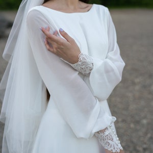 May include: A white wedding dress with long sleeves and a veil. The sleeves feature lace detailing and pearl buttons. The dress has a simple, elegant design.