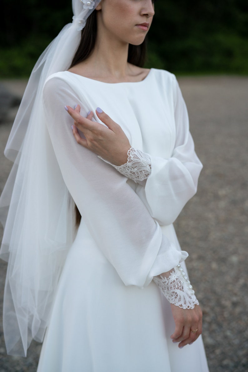 May include: A white wedding dress with long sleeves and a veil. The sleeves feature lace detailing and pearl buttons. The dress has a simple, elegant design.