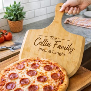 May include: A wooden pizza peel with the text "The Collin Family Pizza & Laughs" is held above a pepperoni pizza. The pizza has a golden crust and is on a wooden cutting board. A small potted plant and tomatoes are in the background.