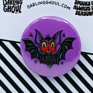 May include: A purple button with a cartoon bat illustration. The bat has a red face, black wings, and is surrounded by stars. The button is on a white card with black and white diagonal stripes and the text "Darling Ghoul". The text "Spooky is always in season!" is also visible.