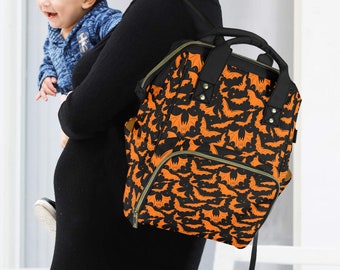 May include: Black and orange bat-patterned backpack with a top handle and pockets. The bag is carried by a person in a black dress. The backpack is designed for carrying baby supplies, ideal for parents.