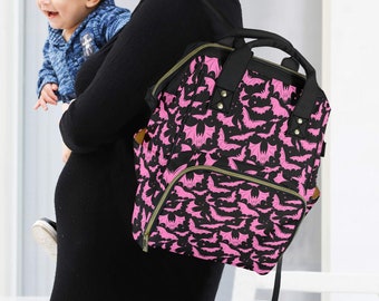 May include: A black and pink bat-patterned backpack with a top handle and multiple pockets. The bag is being carried by a person holding a baby. The backpack has a rectangular shape and is made of a durable material.