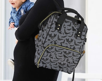 May include: A grey and black bat-patterned nappy bag. The bag features a top handle, a front pocket, and gold-coloured zips. The bag is carried by a person holding a baby.
