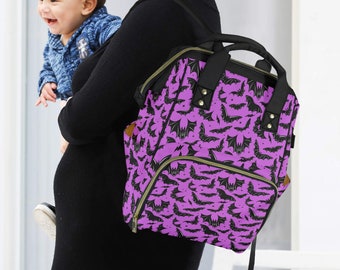 May include: A purple and black bat-patterned nappy bag with black accents. The bag has a top handle, a front pocket, and adjustable straps. The bag is being carried by a person holding a baby.