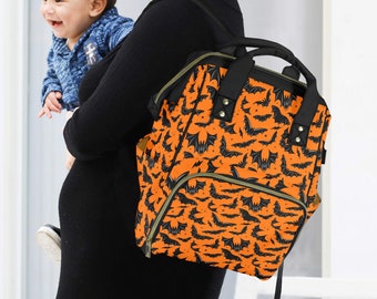 May include: An orange and black backpack with a bat pattern. The backpack has multiple pockets and a top handle. The bag is being carried by a person holding a baby.