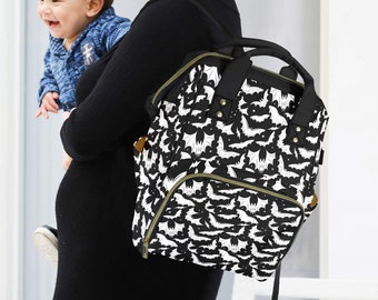 May include: A black and white nappy bag with a bat pattern. The bag has a large main compartment, a front pocket, and two side pockets. The bag is being worn by a person holding a baby.
