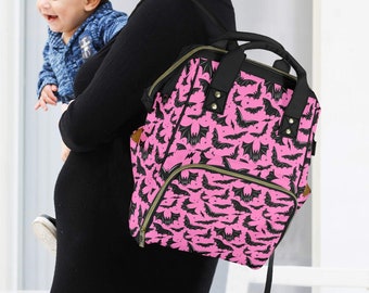 May include: A pink and black bat-print backpack-style changing bag with multiple pockets and a top handle. The bag is carried by a person in a black dress. A baby in a blue jacket is being held.
