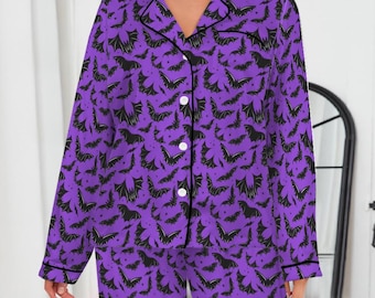 May include: Purple pajama set with a black bat pattern. The long-sleeved top has a button-down front and a notched collar. The pants are long and straight-legged. The set is trimmed with black piping.