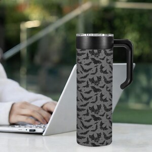May include: A gray travel mug with a black handle and lid. The mug is covered in a pattern of black bats. The mug is cylindrical and appears to be made of stainless steel. The mug is designed to keep beverages hot or cold.