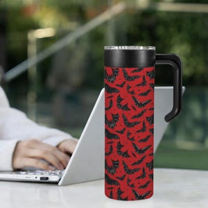 May include: A red travel mug with a black bat pattern. The mug has a black handle, a black lid, and a silver rim. The background is blurred, showing a person using a laptop.