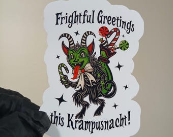 May include: A white sticker with a cartoon Krampus illustration. The green Krampus holds a candy cane and has a red tongue. The sticker reads "Frightful Greetings this Krampusnacht!" in black text with red accents.