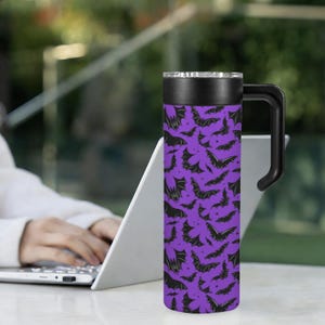 May include: A purple travel mug with a black handle and lid. The mug is covered in a pattern of black bats. The mug is cylindrical and appears to be made of stainless steel. The handle is curved and attached to the side of the mug.