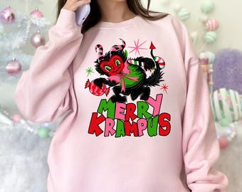 May include: A light pink sweatshirt featuring a black Krampus graphic with a green and pink bow, holding candy. The text "MERRY KRAMPUS" is in green and red. The sweatshirt is a festive holiday design.