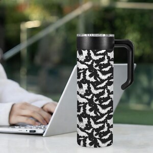 May include: A black and white travel mug featuring a repeating pattern of bats. The mug has a black handle, a black lid, and a silver rim. The mug is cylindrical and appears to be made of stainless steel.