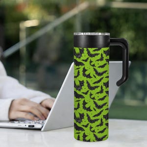 May include: A green travel mug with a black handle and lid. The mug is covered in a pattern of black bats. The mug is cylindrical and appears to be made of stainless steel. The mug is designed for hot or cold beverages.