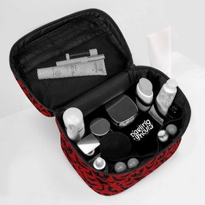 May include: Red and black travel makeup bag with a bat print and the text "Darling Ghouls" on the front. The bag is open and filled with various cosmetics and toiletries.