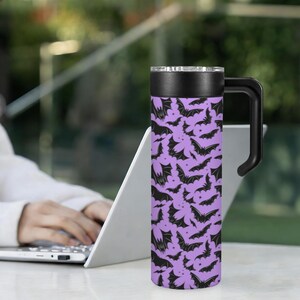 May include: A purple travel mug with a black handle and lid. The mug is covered in a pattern of black bats. The mug is cylindrical and appears to be made of stainless steel. The mug is designed to keep beverages hot or cold.