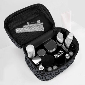 May include: Black travel makeup bag with a bat print design. The bag is open and filled with various cosmetics and toiletries, including a white tube, a black jar, and a silver bottle. The bag has a mesh pocket on the inside.