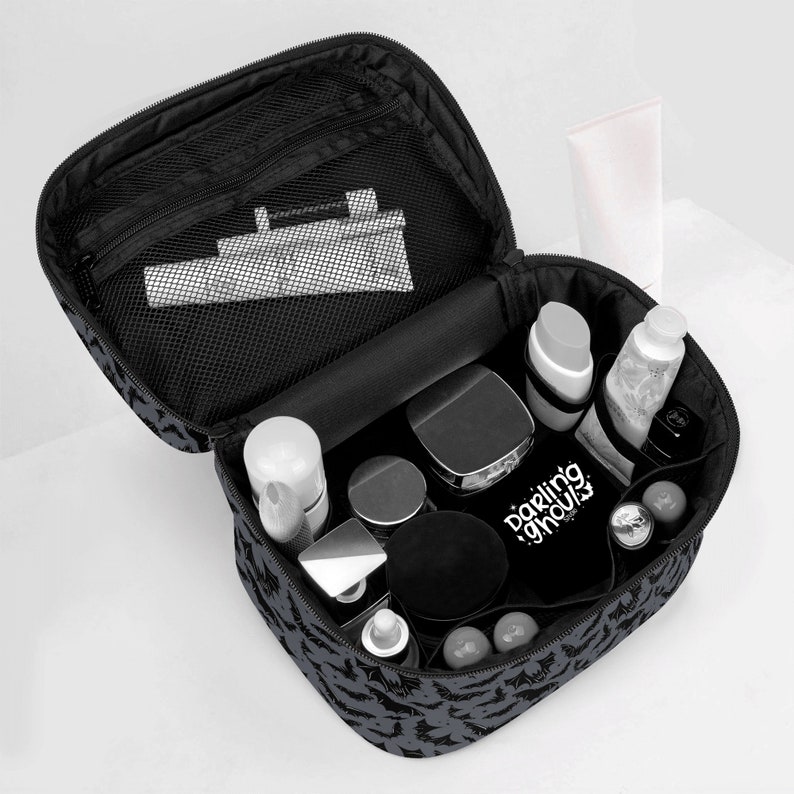 May include: Black travel makeup bag with a bat print design. The bag is open and filled with various cosmetics and toiletries, including a white tube, a black jar, and a silver bottle. The bag has a mesh pocket on the inside.