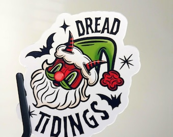 May include: A die-cut sticker featuring a red-faced, horned character with a white beard wearing a green hat. The text DREAD TIDINGS is printed in black, accompanied by black bat silhouettes and star icons on a white background.