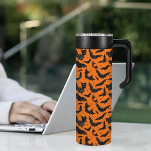 May include: A tall, orange travel mug with a black handle and lid. The mug is covered in a pattern of black bats. The mug is cylindrical and appears to be made of stainless steel. The mug is sitting on a white surface.